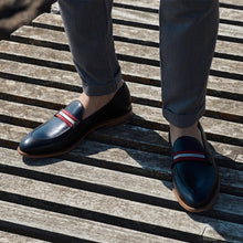 Load image into Gallery viewer, A lifestyle shot of black calf web stripe loafers worn with grey trousers, capturing the shoes' elegant integration into a smart-casual ensemble, set on a wooden deck.
