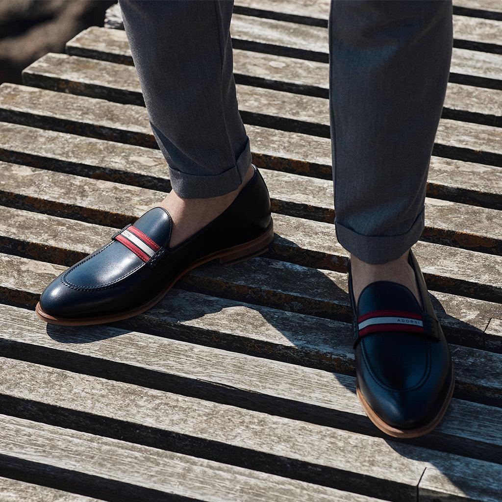 A lifestyle shot of black calf web stripe loafers worn with grey trousers, capturing the shoes' elegant integration into a smart-casual ensemble, set on a wooden deck.