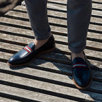 A lifestyle shot of black calf web stripe loafers worn with grey trousers, capturing the shoes' elegant integration into a smart-casual ensemble, set on a wooden deck.