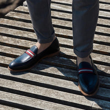 Load image into Gallery viewer, A lifestyle shot of black calf web stripe loafers worn with grey trousers, capturing the shoes&#39; elegant integration into a smart-casual ensemble, set on a wooden deck.
