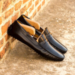Lifestyle shot of black nappa leather penny driving loafers with wooden shoe trees inserted, casually leaning against a rustic brick wall.