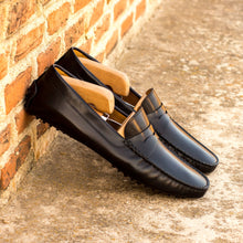 Load image into Gallery viewer, Lifestyle shot of black nappa leather penny driving loafers with wooden shoe trees inserted, casually leaning against a rustic brick wall.
