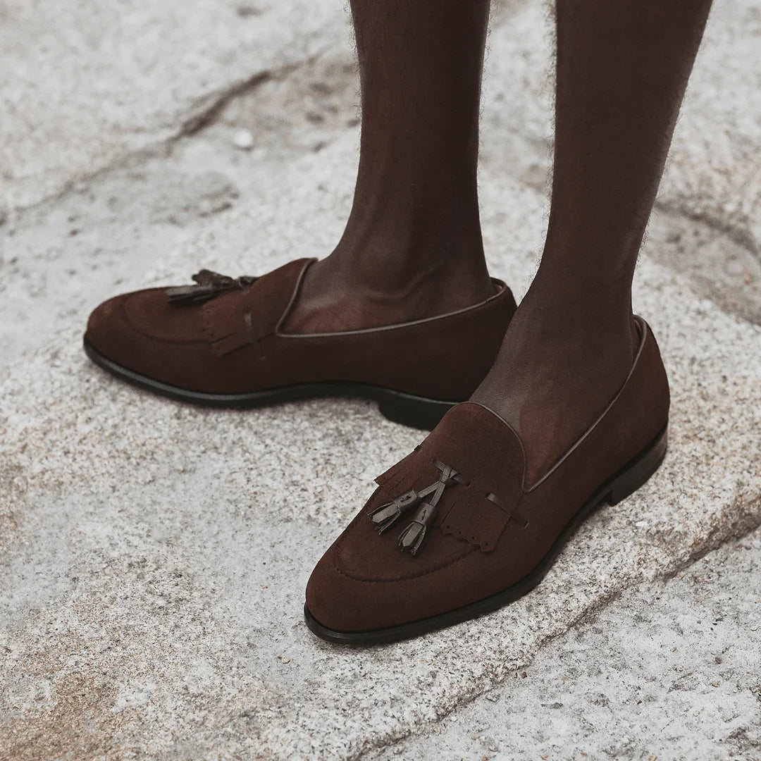 A lifestyle shot showcasing a person wearing the brown suede kiltie tassel loafers on a textured pavement, highlighting the shoe's casual elegance.