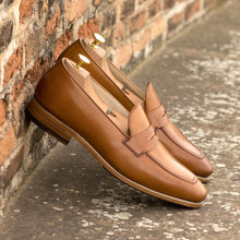 Load image into Gallery viewer, Lifestyle shot of handcrafted cognac shell cordovan penny loafers casually placed against a rustic brick wall, exuding a sense of timeless style.
