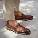 Lifestyle image of someone wearing burnished medium brown calf plain loafers paired with beige trousers, captured on a textured grey sidewalk.