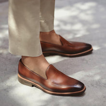 Load image into Gallery viewer, Lifestyle image of someone wearing burnished medium brown calf plain loafers paired with beige trousers, captured on a textured grey sidewalk.
