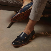 Load image into Gallery viewer, Casual setting with a model wearing the navy fringe penny loafers, paired with light grey trousers, illustrating the shoe&#39;s versatility and elegance.
