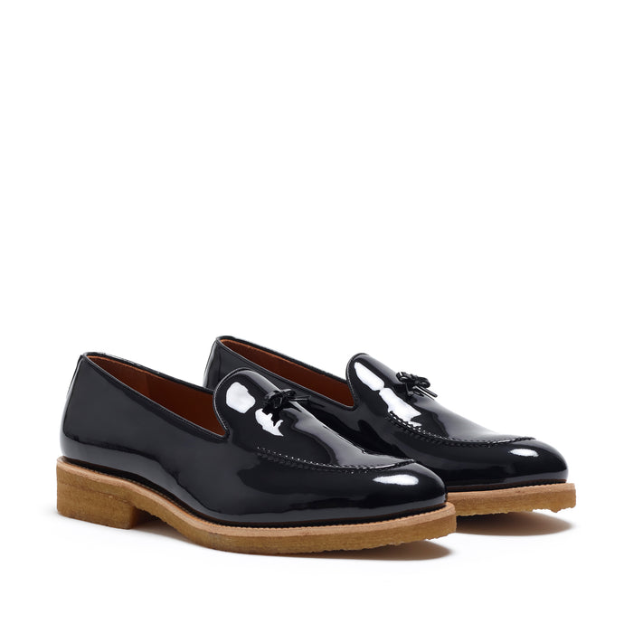 A pair of glossy patent black Belgian loafers with a distinctive bow, set against a white background, highlighting the mirror-like finish.