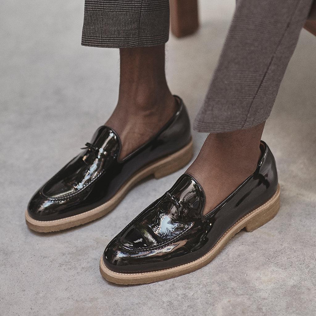 A lifestyle shot of a person wearing patent black Belgian loafers with grey trousers, demonstrating the shoe's elegance for smart casual attire.