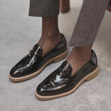 A lifestyle shot of a person wearing patent black Belgian loafers with grey trousers, demonstrating the shoe's elegance for smart casual attire.